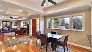 A spacious living room with hardwood floors, a dining area with a table and chairs, and large windows offering a view of trees and a house. - Two Sweet by Avantstay Walk to Downtown + Private Patio Historic Paso Robles Wine Country (Paso Robles)