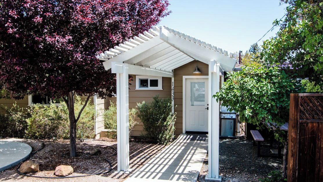 A covered patio area with a pergola, a door, and a small garden with a tree and shrubs.