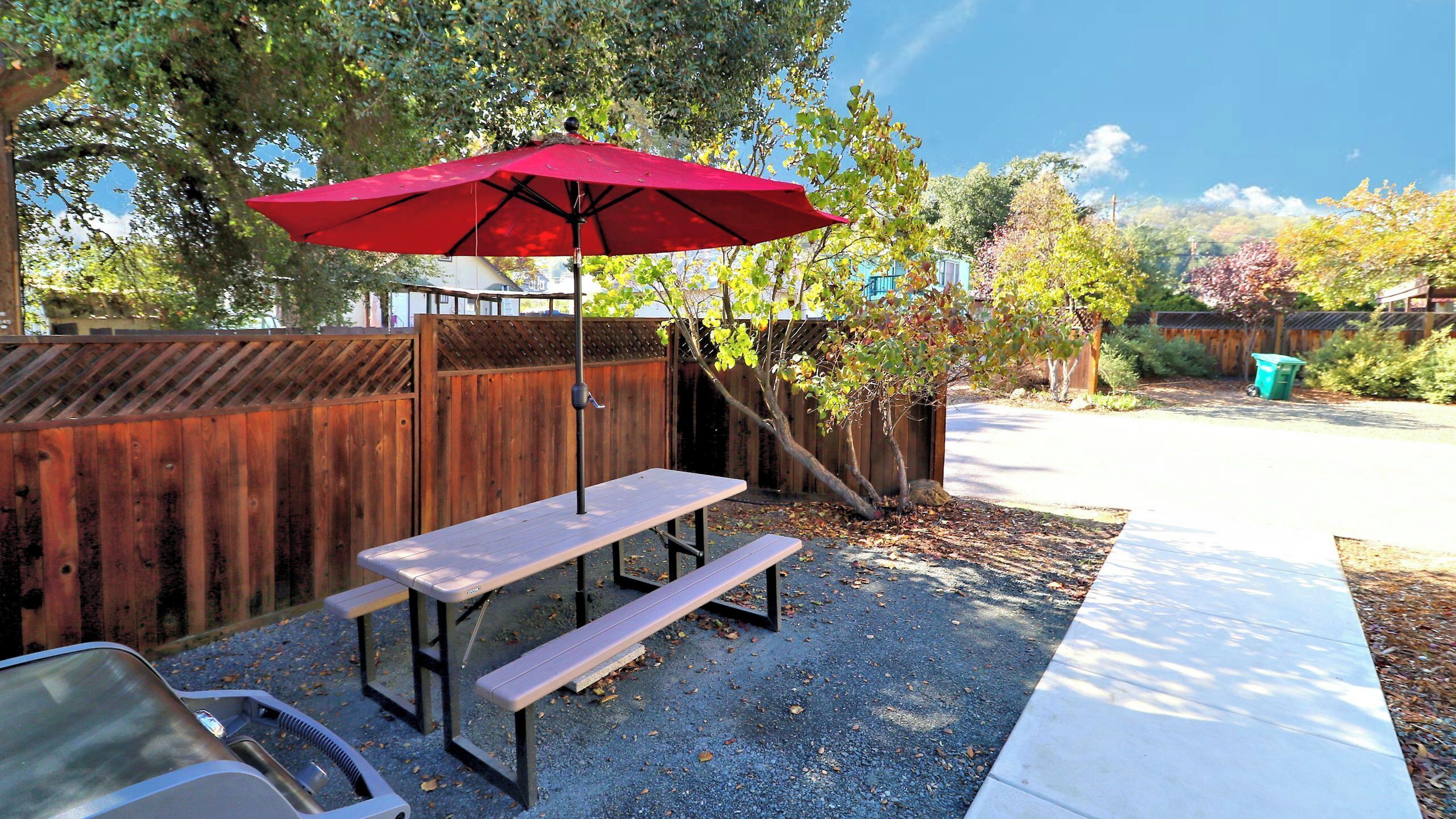 A picnic table with a red umbrella, a wooden fence, and a gravel ground.