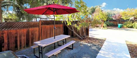A picnic table with a red umbrella, a wooden fence, and a gravel ground.