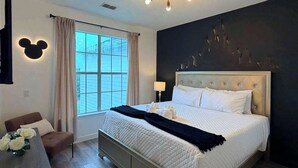 Ein Schlafzimmer mit Bett, Nachttisch, Stuhl, einem Fenster mit Vorhängen, einer Wanddekoration mit Mickey Mouse und einer Deckenleuchte.