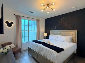 A bedroom with a bed, a bedside table, a chair, a window with curtains, a wall-mounted Mickey Mouse decoration, and a ceiling light fixture. - Secret Lake Resort Balcony Retreat (Kissimmee)
