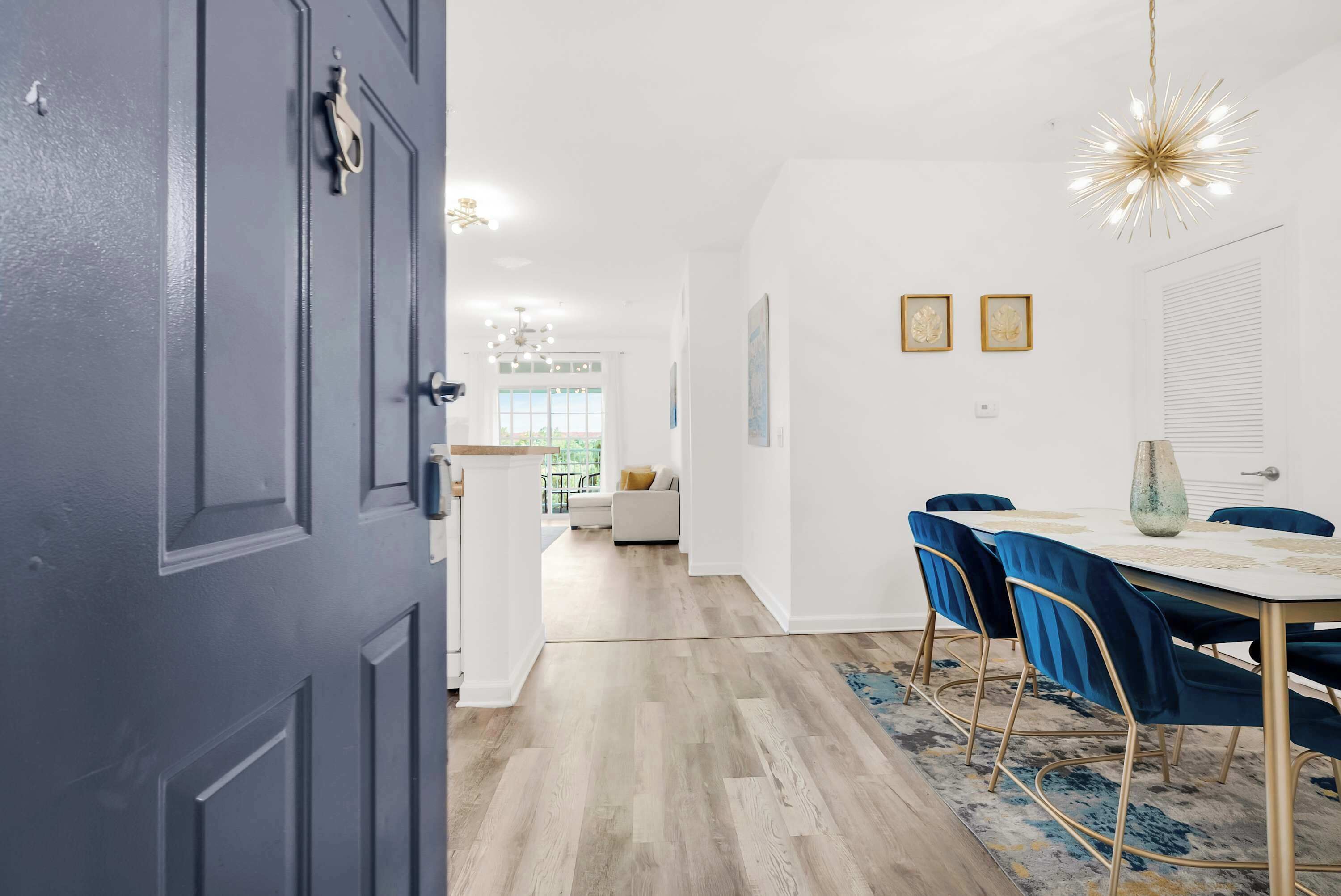 Un interior moderno con comedor que presenta una mesa blanca y sillas azules, una araña y vista al salón.
