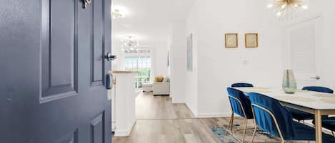 A modern interior with a dining area featuring a white table and blue chairs, a chandelier, and a view into the living room.