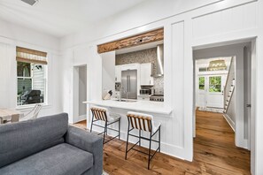 A modern kitchen with white cabinetry, a stainless steel refrigerator, and a wooden beam ceiling. There are two bar stools and a gray sofa in the living area. - Next to Camp Watercolor and Watercolor Publix (Santa Rosa Beach)