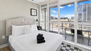 Habitación de hotel con una cama grande, vistas de la ciudad a través de ventanales del suelo al techo y un balcón con mobiliario exterior.
