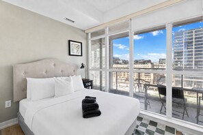 A hotel room with a large bed, a view of the city through floor-to-ceiling windows, and a balcony with outdoor seating.