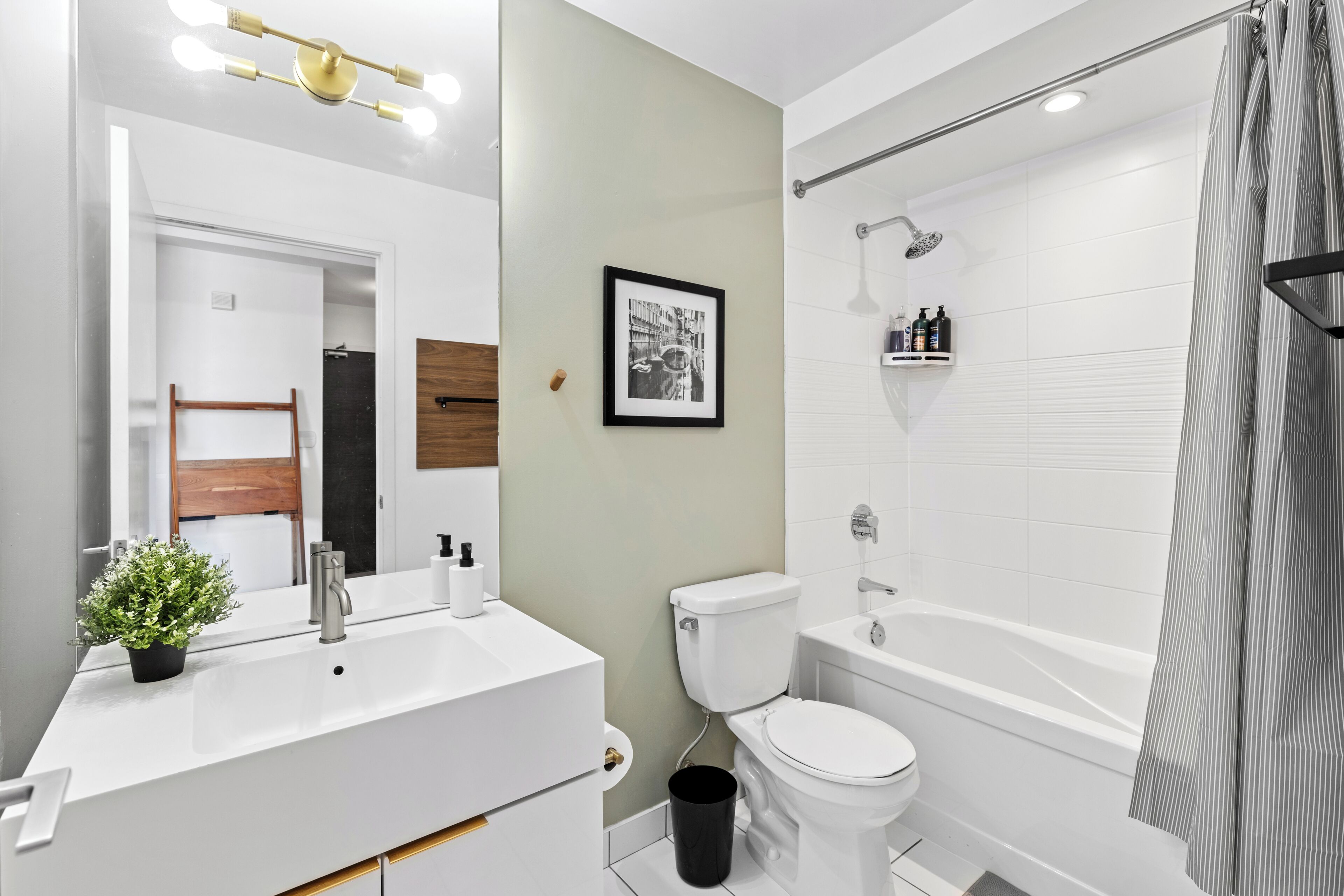 A modern bathroom with a white sink, toilet, and bathtub. There is a framed picture on the wall and a small plant on the sink.