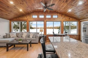 A spacious living area with a wooden ceiling, a large window overlooking a body of water, and a kitchen with modern appliances. - Breakaway Lake House (Northwood)