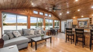 A spacious living area with a wooden ceiling, a sofa, a coffee table, and a kitchen area with a bar. - Breakaway Lake House (Northwood)
