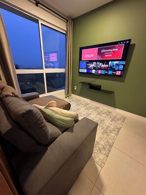 A living room with a large window, a flat-screen TV mounted on the wall, and a sofa with cushions. - Fully furnished apartment | 1 bedroom | Garage | Dom Bosco - Itajaí (Itajaí)
