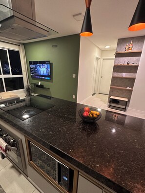 A modern kitchen with a black granite countertop, a bowl of fruit, a built-in oven, and a television mounted on the wall. - Fully furnished apartment | 1 bedroom | Garage | Dom Bosco - Itajaí (Itajaí)