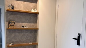A hallway with a white door, a wooden bench, and three wooden shelves against a textured wall. - Fully furnished apartment | 1 bedroom | Garage | Dom Bosco - Itajaí (Itajaí)