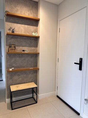 A hallway with a white door, a wooden bench, and three wooden shelves against a textured wall. - Fully furnished apartment | 1 bedroom | Garage | Dom Bosco - Itajaí (Itajaí)