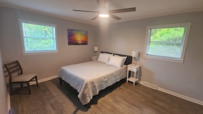 A bedroom with a bed, two bedside tables, a chair, a ceiling fan, and two windows with blinds.