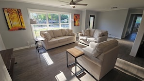 A living room with a sofa, armchair, and coffee table, large windows with a view, and a ceiling fan.