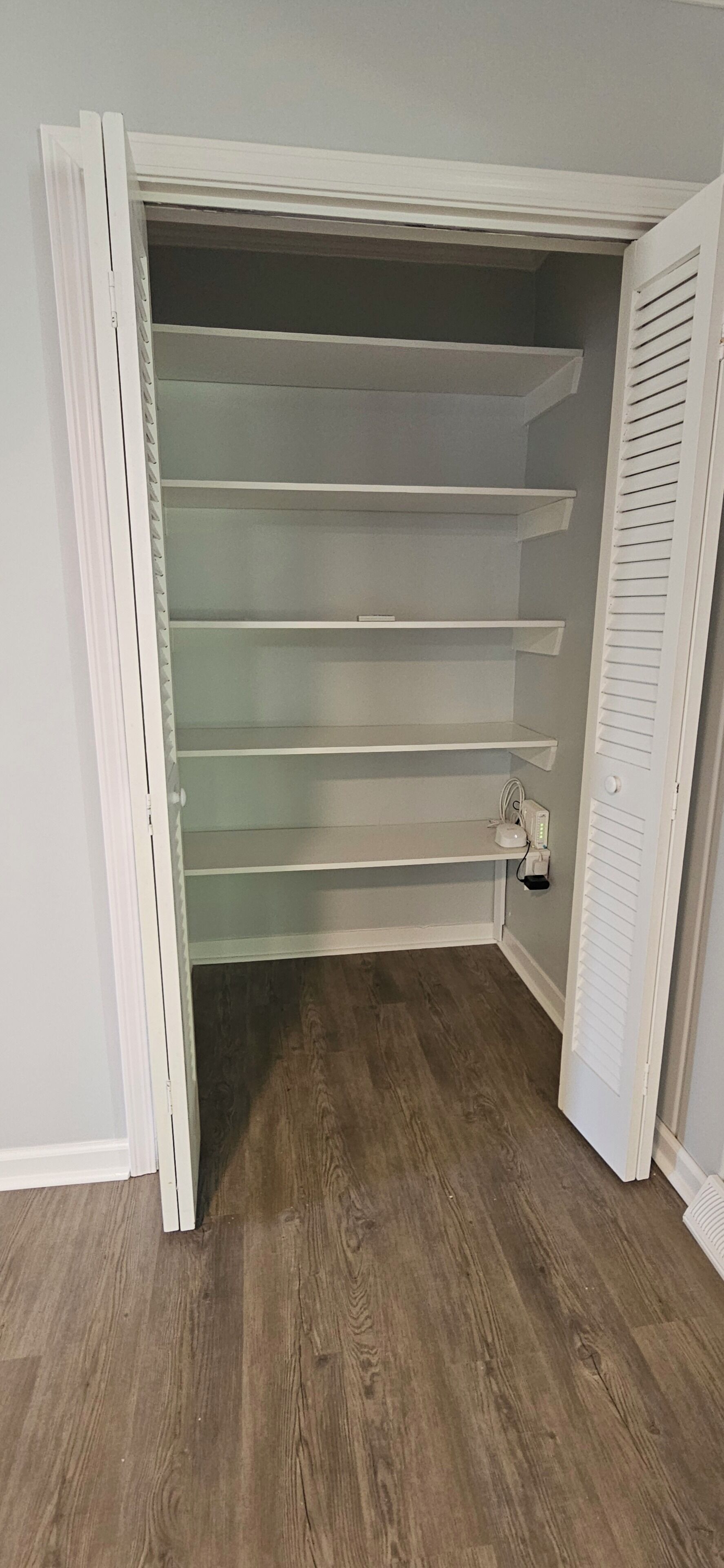 A walk-in closet with open doors, revealing multiple shelves and a power outlet on the floor.