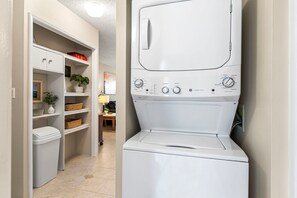 A laundry room with a front-loading washing machine, shelves with baskets, and a trash bin. - 🏖️Alii Villas 317 | Sunset Views | Remodeled | Pool & A/C (Kailua Kona)