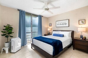 A hotel room with a bed, a ceiling fan, a window with curtains, a wall-mounted picture, and a bedside lamp. - 🏖️Alii Villas 317 | Sunset Views | Remodeled | Pool & A/C (Kailua Kona)
