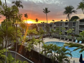 A pool area with lounge chairs and palm trees, surrounded by a building with multiple balconies. - 🏖️Alii Villas 317 | Sunset Views | Remodeled | Pool & A/C (Kailua Kona)