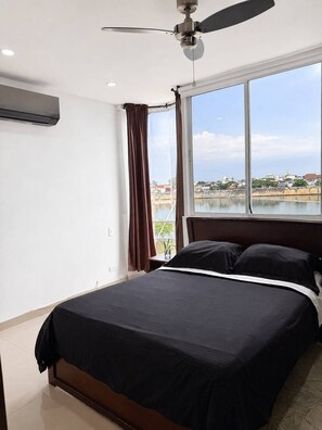 A bedroom with a bed, a ceiling fan, a window with a view of a body of water and buildings, and an air conditioning unit on the wall. - 2BR/2BA Bay View Apartment | 500 Mbps WiFi | Near Getsemaní (Cartagena)