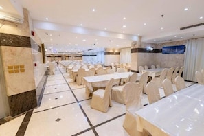 Un salón de banquetes con varias mesas redondas y sillas, piso de mármol y una pantalla grande en la pared.