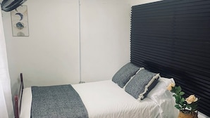 A single bed with a white mattress, grey and white bedding, a black headboard, a small bedside table with a plant, and a ceiling fan.