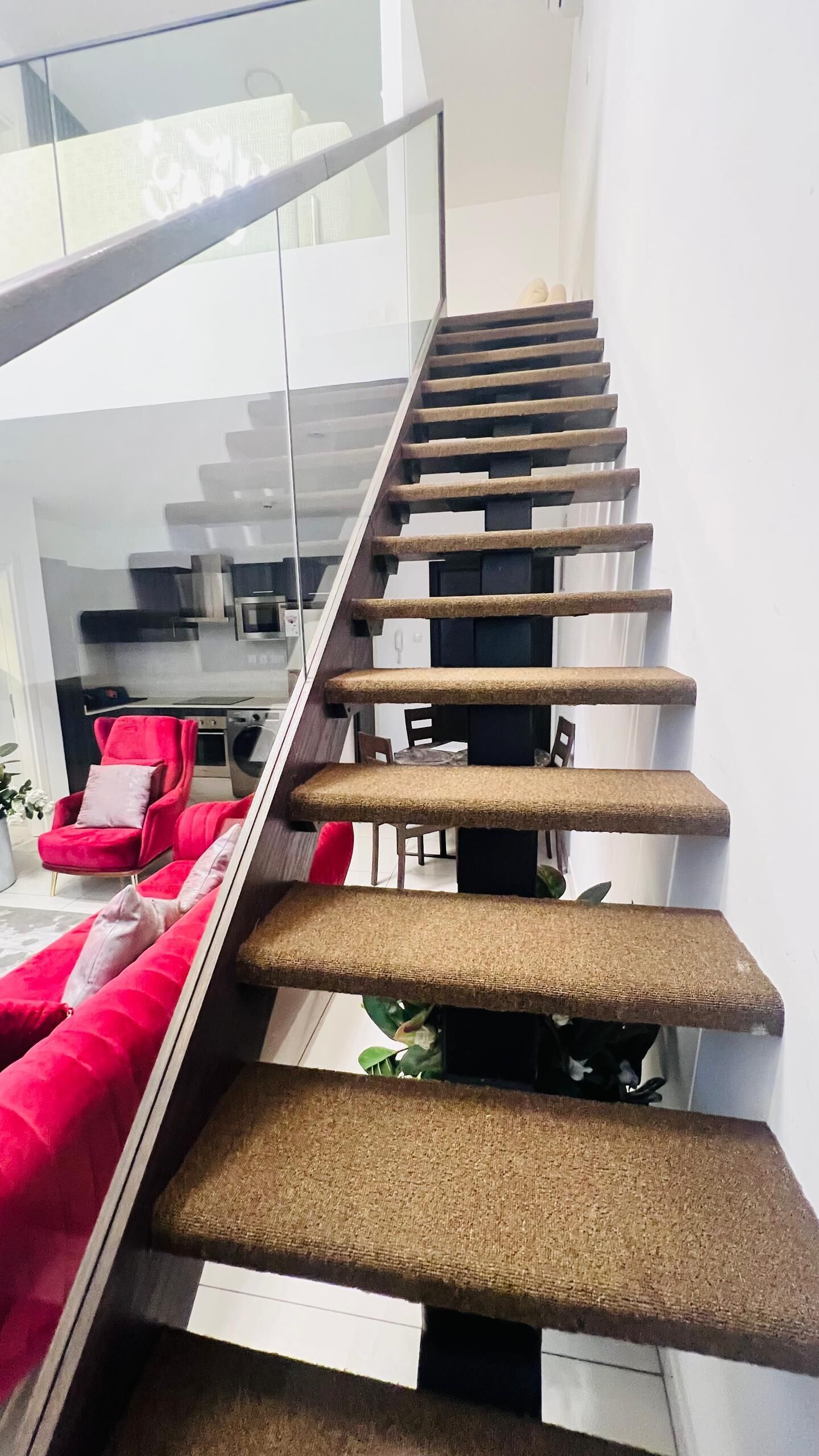 Una escalera moderna con barandilla de vidrio y una silla roja al fondo.