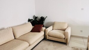 A living room with a beige sofa, a patterned armchair, a small table, and a potted plant. - EBS PROPERTIES (Accra)