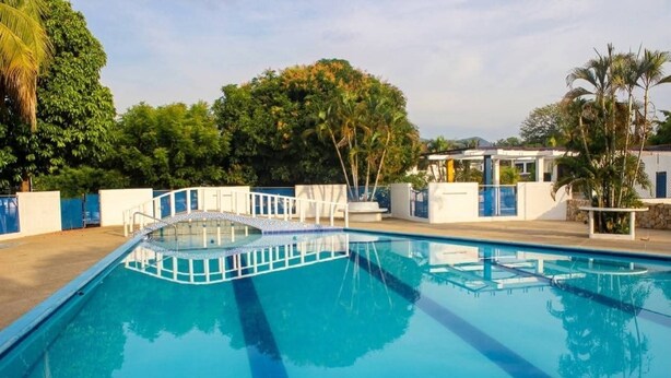 Una piscina con terraza blanca, rodeada de vegetación y un edificio con una combinación de colores azul y blanco.