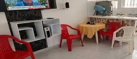 A room with a television mounted on the wall, a kitchen area with a refrigerator, and a dining table with chairs.