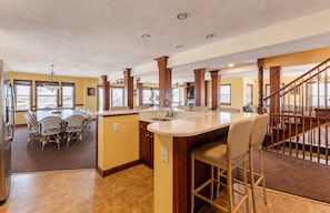 A spacious kitchen with a central island, a dining area with wicker chairs, and a staircase leading to an upper level. - The Sea Spirit-KDH MP 7, Oceanside, Elevator, Pool, Hot Tub, Pets OK, YR Flex Stays (Kill Devil Hills)