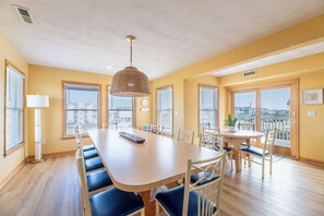 A dining area with a long table, chairs, a hanging light fixture, and large windows with blinds. - Captains Choice-KDH MP 7, Oceanside, Pool, Hot Tub, Pets OK, YR Flex Stays (Kill Devil Hills)