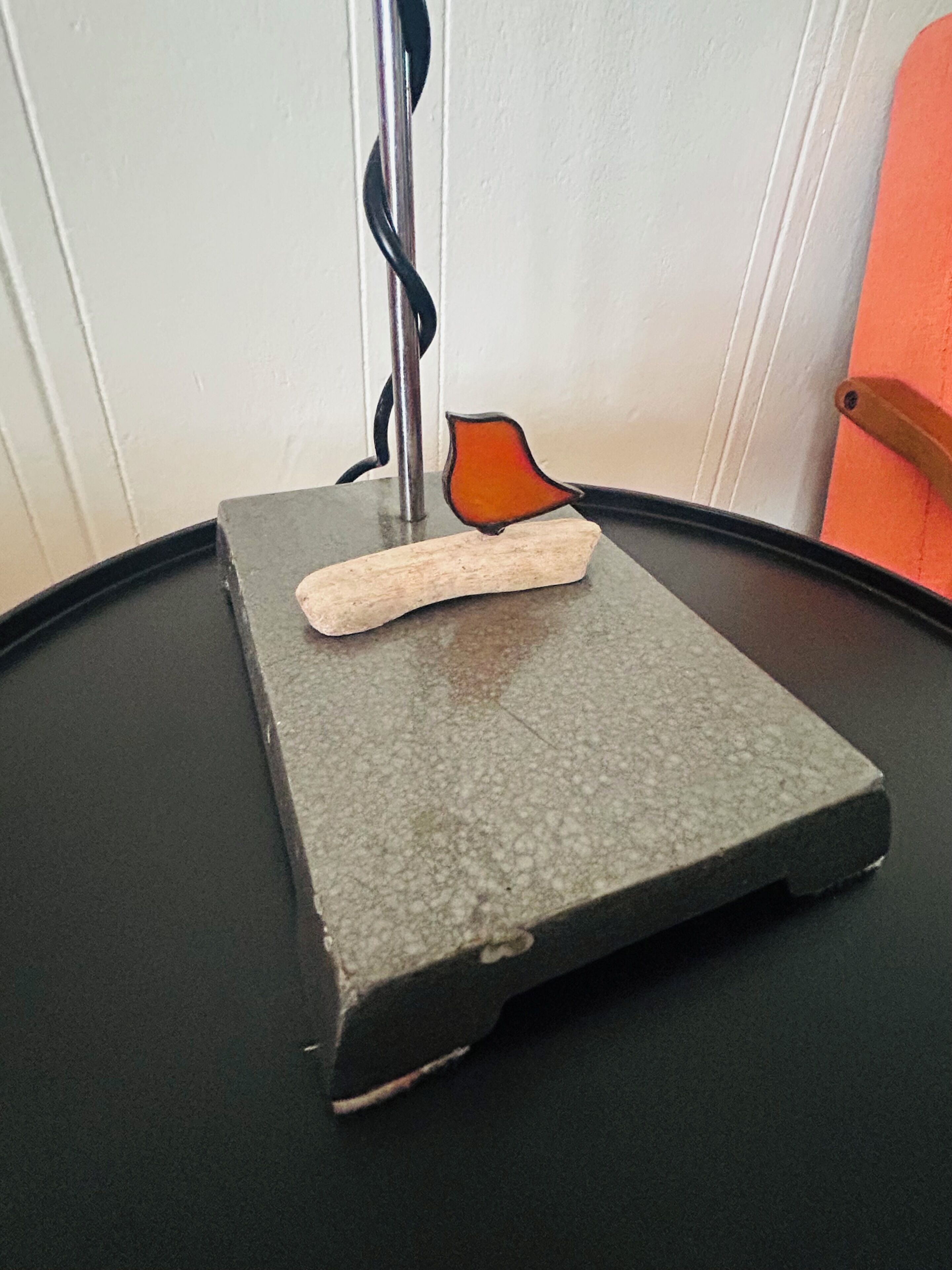 Ein kleiner, quadratischer Tisch mit einer strukturierten Oberfläche, darauf ein weißes Objekt, vermutlich ein Buch oder eine Schachtel, und ein schwarzes Kabel, das vertikal verläuft.