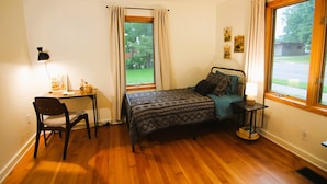 A bedroom with a bed, a desk with a chair, a table with a lamp, and a window with curtains. - Brand new listing, be the first to stay at this charming downtown Crosby home. (Crosby)