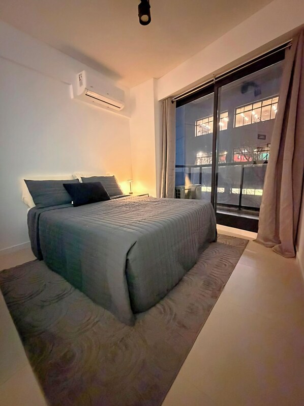 Ein Schlafzimmer mit einem Bett, einem Nachttisch, einer Klimaanlage und einem großen Fenster mit Vorhängen.