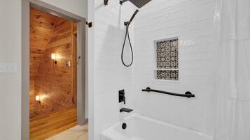 Un bagno moderno con soffitto in legno, pareti rivestite di piastrelle bianche, una vasca, una doccia con soffione manuale e un piccolo pannello decorativo in piastrelle.