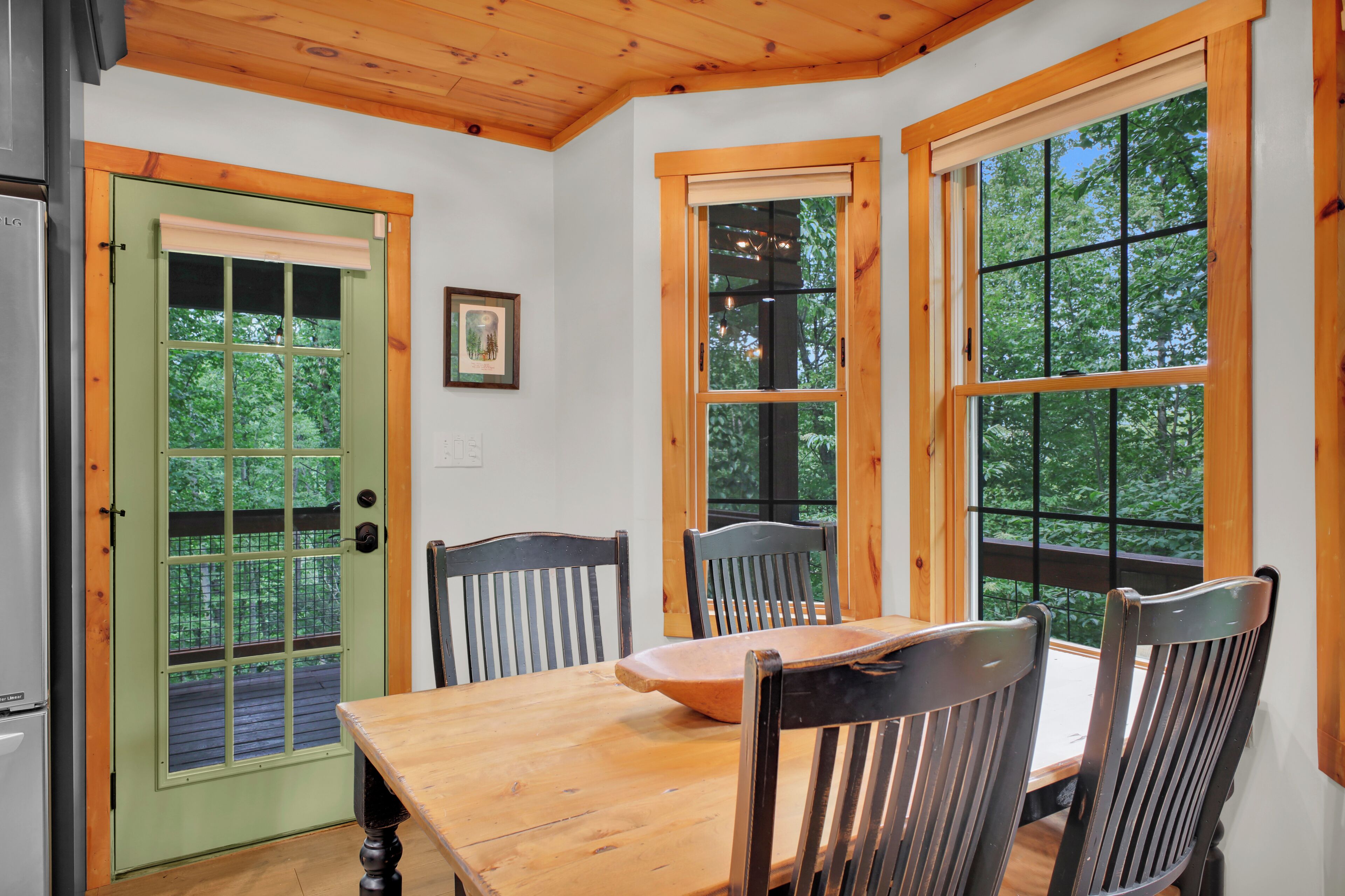 Un comedor con una mesa y sillas de madera, una puerta verde y ventanas grandes con vista a árboles.