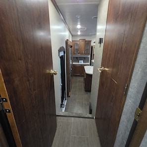 A narrow hallway with wooden doors leading to a kitchen area with cabinets and a countertop.