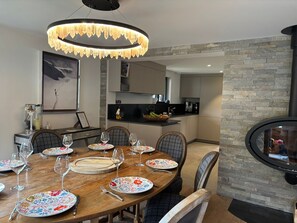 Un comedor con una mesa de madera dispuesta para seis personas, rodeada de sillas. Un candelabro moderno cuelga encima, y se observa una pared de piedra al fondo.