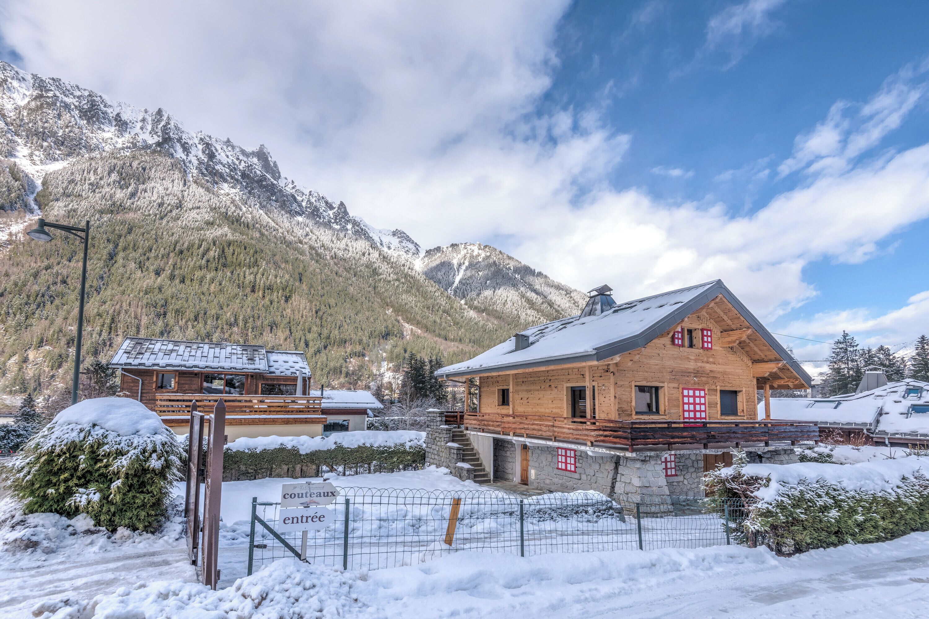 A wooden chalet with a stone foundation, a wooden deck, and a view of snow-covered mountains.