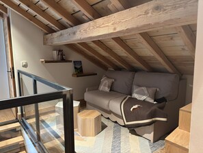 Une chambre mansardée chaleureuse, avec un plafond en pente, un canapé gris agrémenté de coussins décoratifs, une petite table d’appoint en bois et une balustrade en verre donnant sur un balcon.