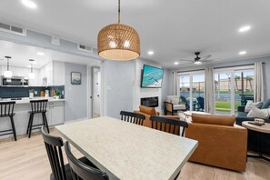A modern kitchen with white cabinets, a dining area with a table and chairs, and a living room with a sofa and a TV. - Ground Floor Condo | 2 King Suites | Beach (Panama City Beach)