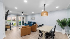 A modern living room with a dining area, a ceiling fan, and a large window overlooking a tennis court. - Ground Floor Condo | 2 King Suites | Beach (Panama City Beach)