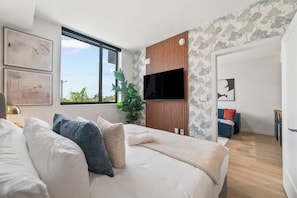 A modern bedroom with a large bed, a TV mounted on a wooden panel wall, and a view of the outdoors through a large window.