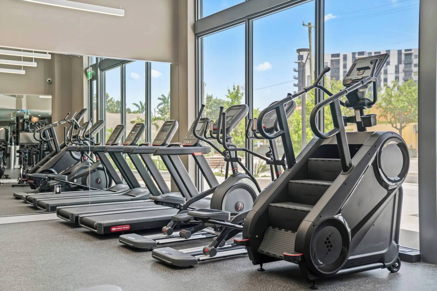 A row of treadmills in a gym with large windows and a view of buildings outside.