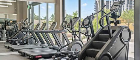 A row of treadmills in a gym with large windows and a view of buildings outside.