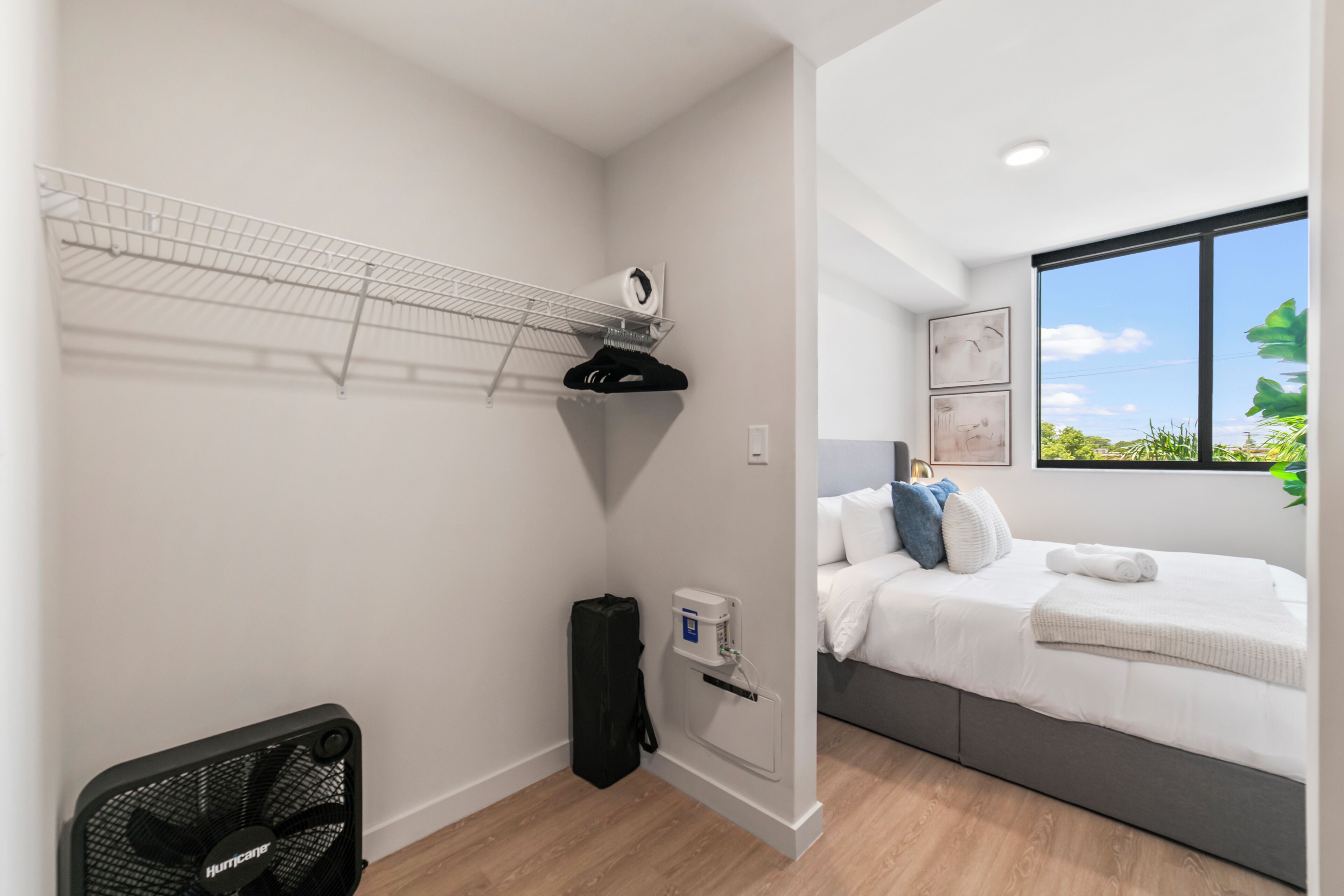 Ein Schlafzimmer mit Bett, Nachttisch, einer an der Wand befestigten Ablage mit Ventilator und einem Fenster mit Blick auf Grünflächen.