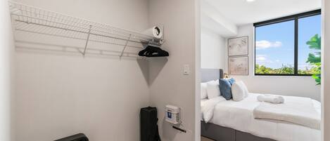 A bedroom with a bed, a bedside table, a wall-mounted shelf with a fan, and a window with a view of greenery.