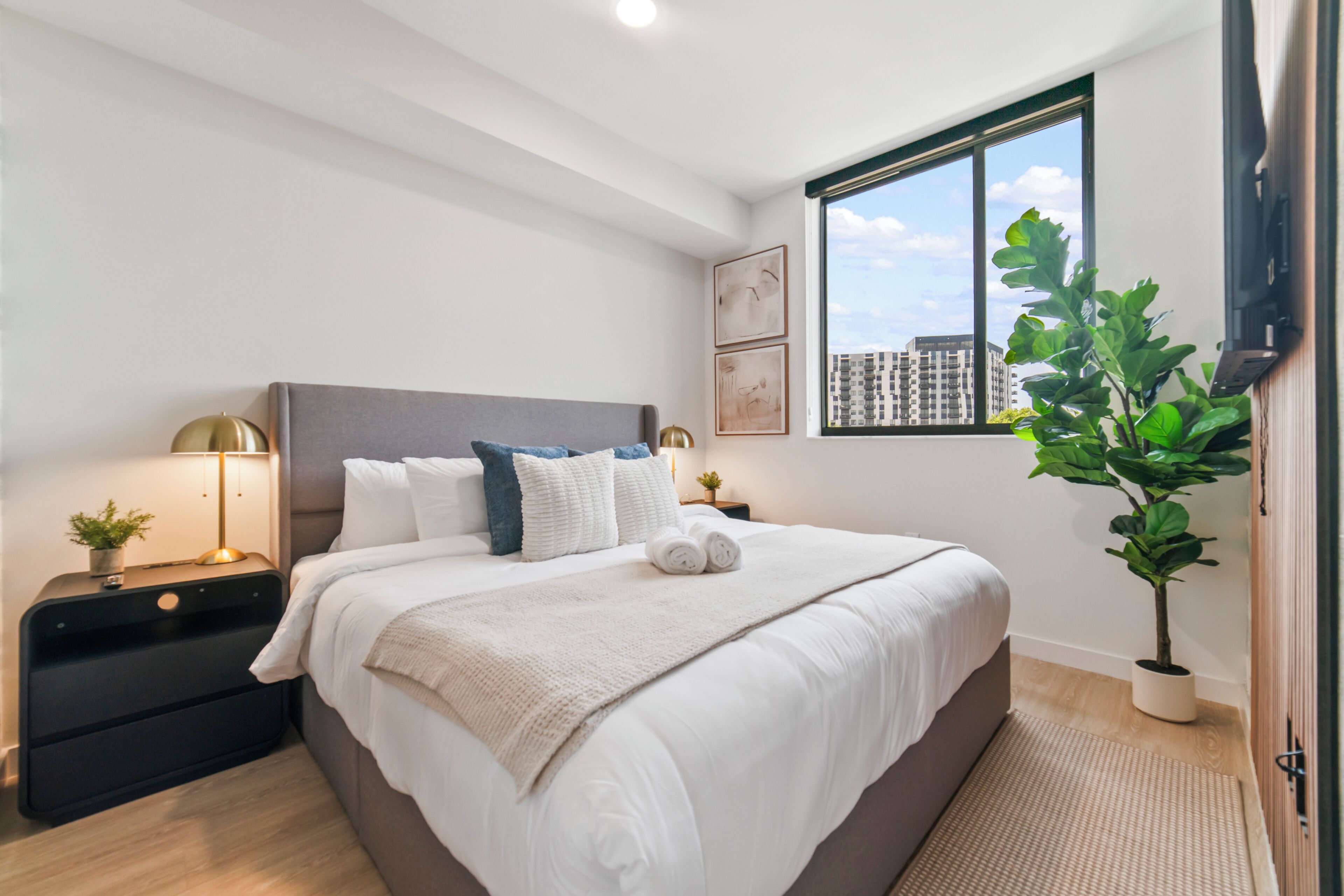 Un dormitorio moderno con una cama grande, mesitas de noche, una planta en maceta y un ventanal con vistas a la ciudad.